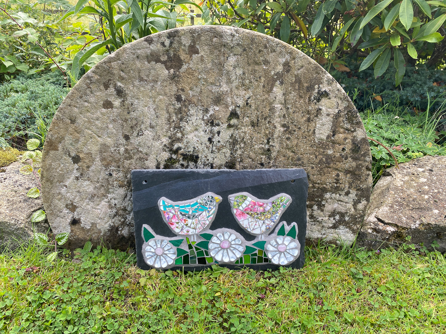 A rectangular black slate wall plaque featuring two colorful mosaic birds, placed outdoors on grass in front of a large weathered stone.