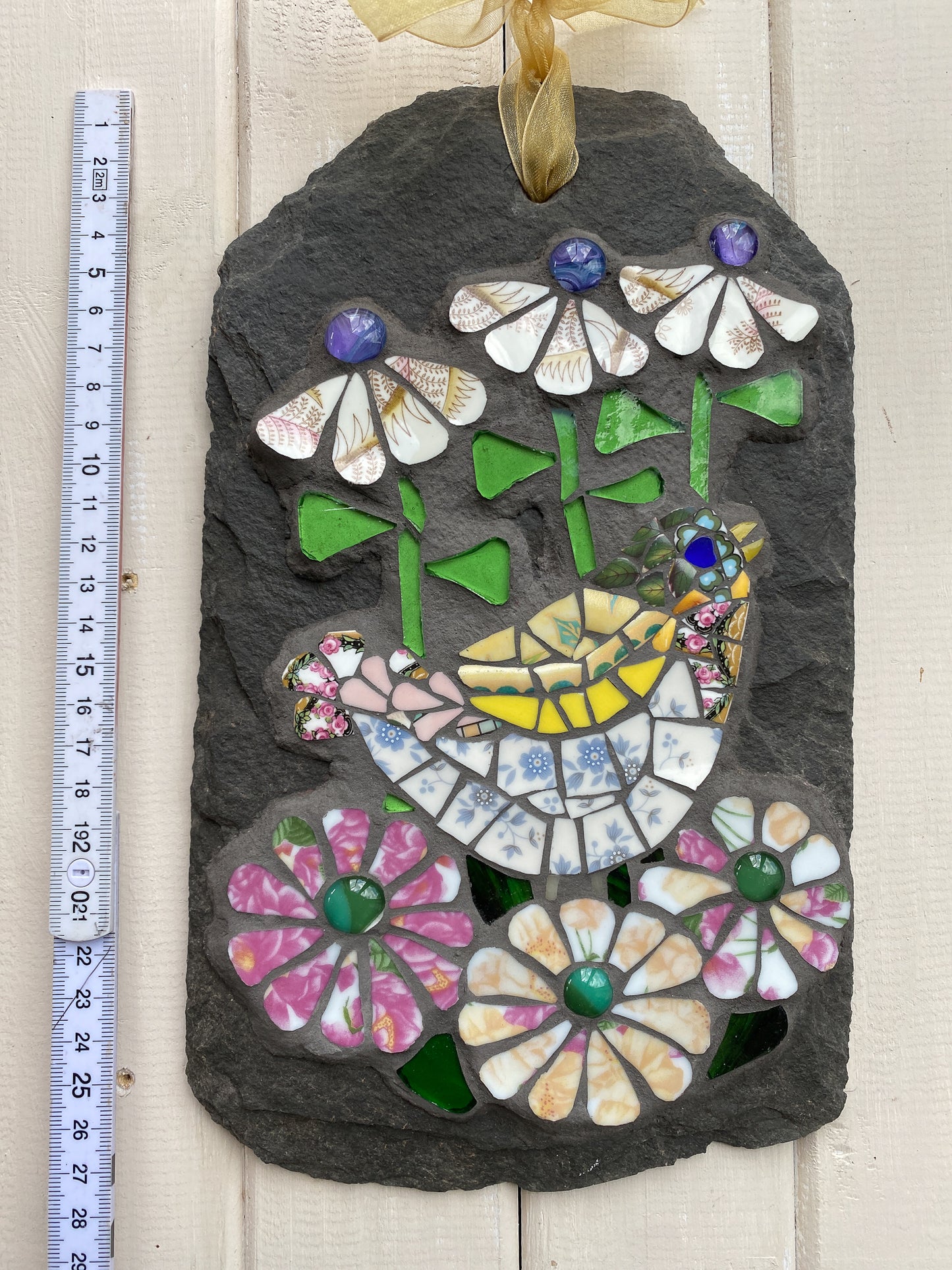 A mosaic garden stone ornament featuring a yellow bird and colorful flowers, shown next to a 2-foot measuring tape on a light wood background.