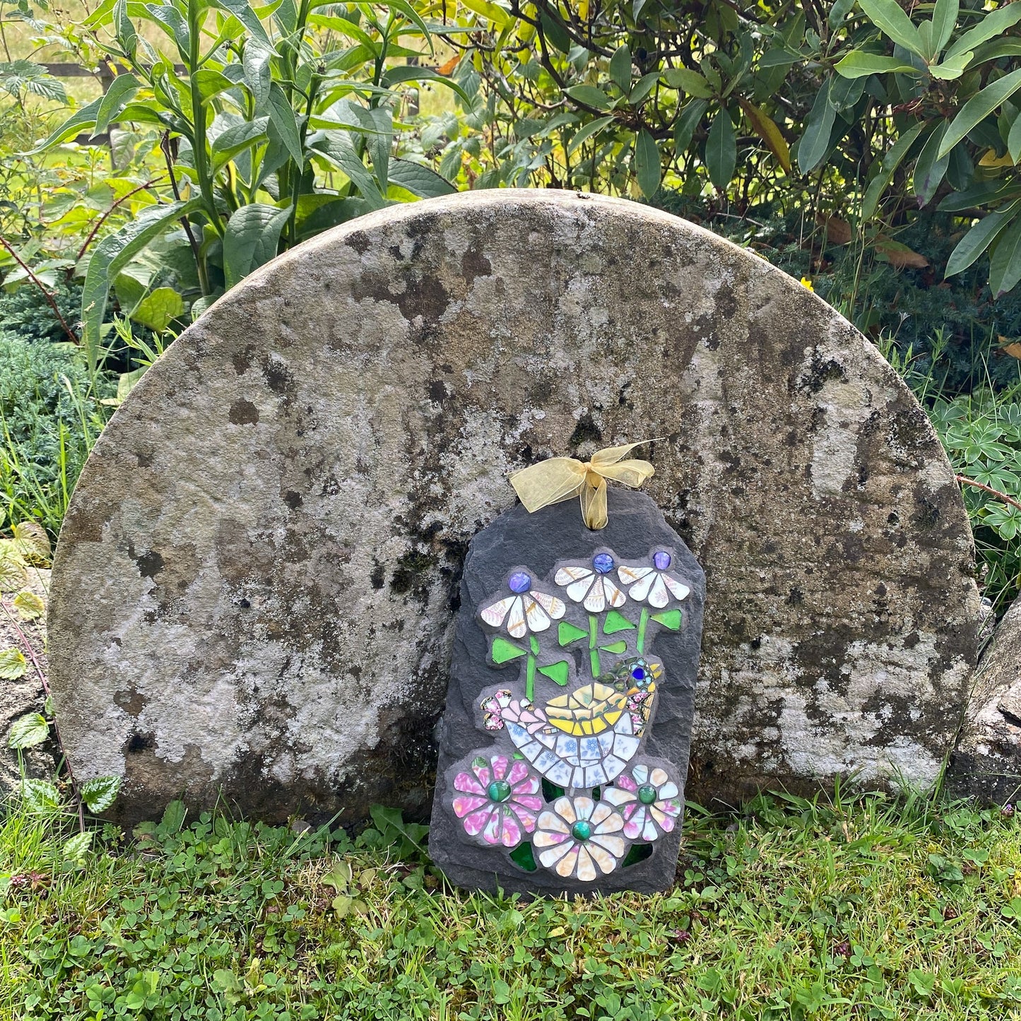A floral mosaic with bird on it, made on slate, standing in front of a large weathered stone slab outdoors on grass with green plants behind it.