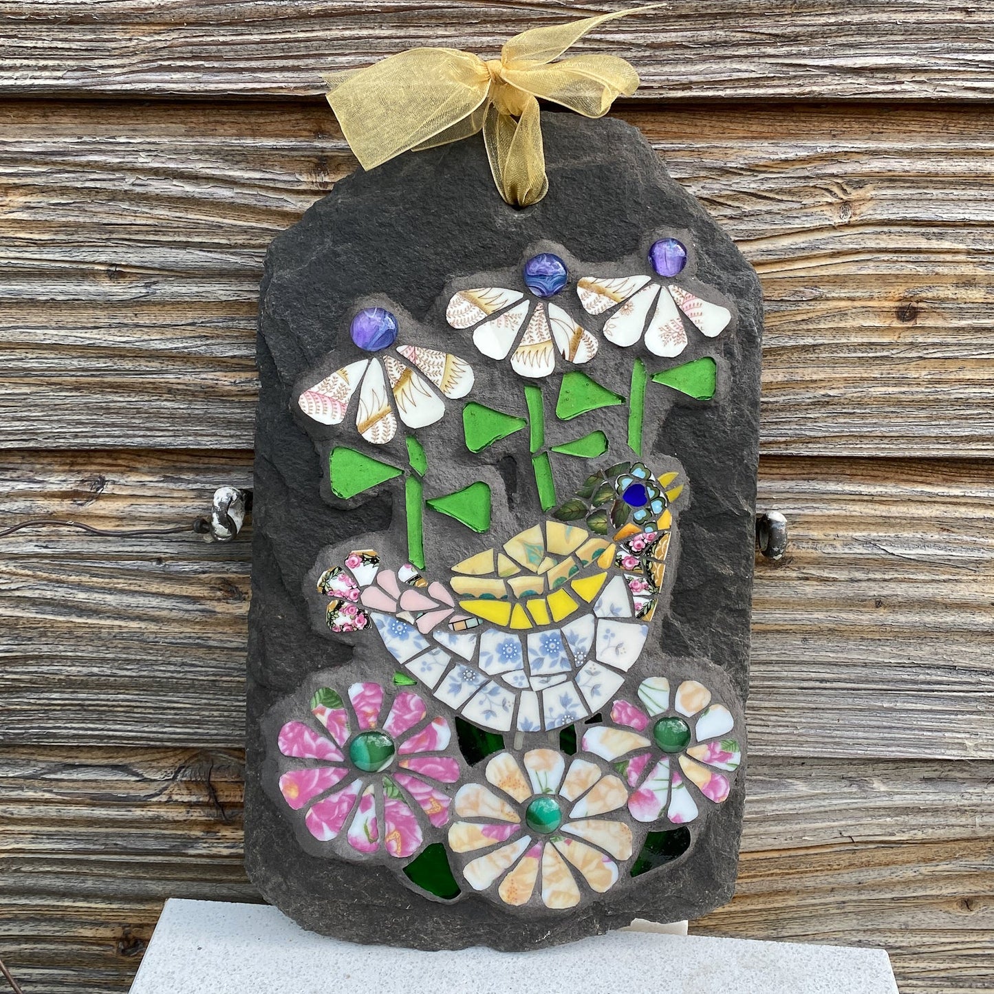 A decorative hanging plaque made from a dark slate with colorful mosaic flowers, leaves, and a small bird, tied at the top with a sheer gold ribbon. The plaque is photographed against a rustic weathered wood plank background.