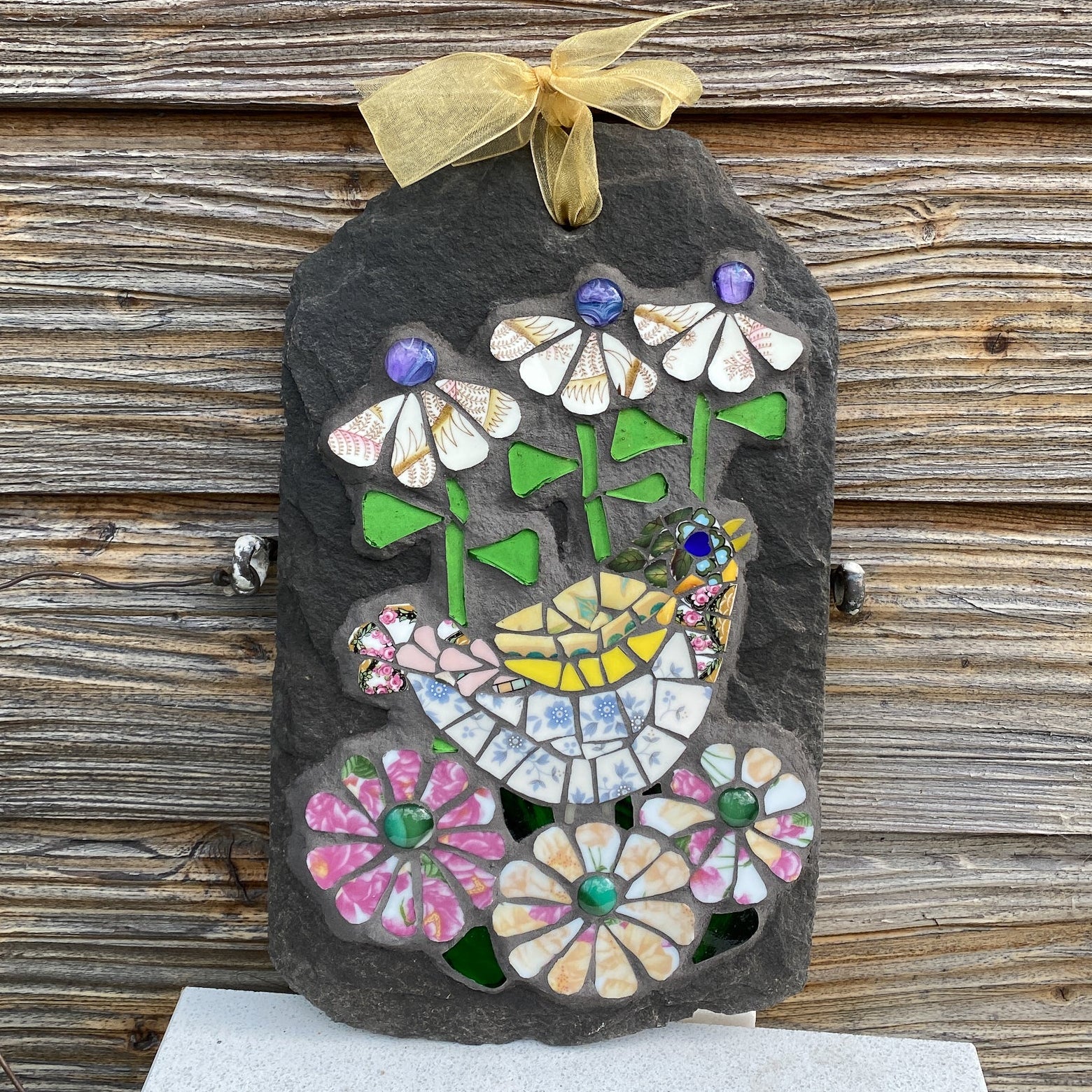 A decorative hanging plaque made from a dark slate with colorful mosaic flowers, leaves, and a small bird, tied at the top with a sheer gold ribbon. The plaque is photographed against a rustic weathered wood plank background.