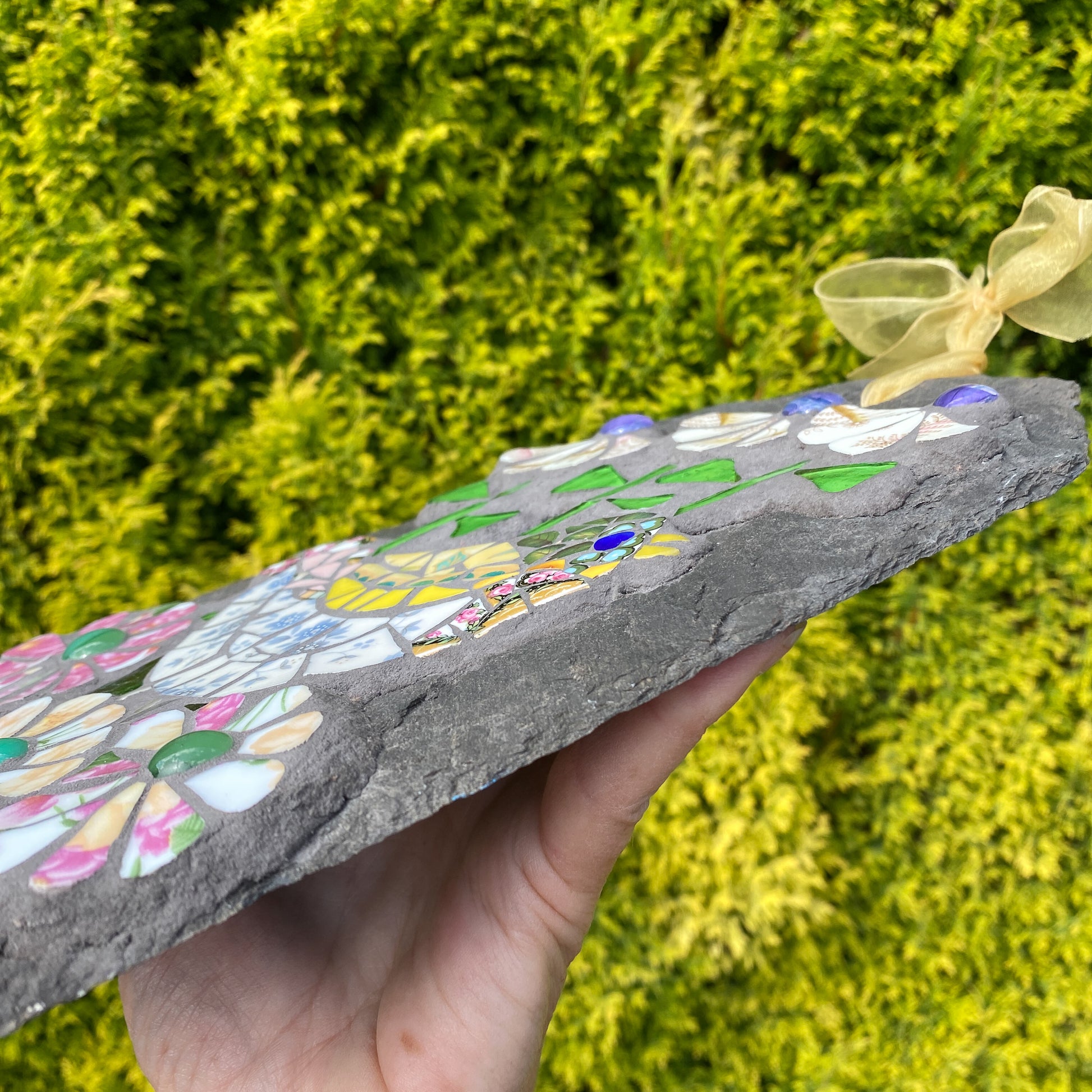 A hand holds a rectangular slate plaque decorated with colorful mosaic flower and butterfly designs, set against a blurred background of green foliage.