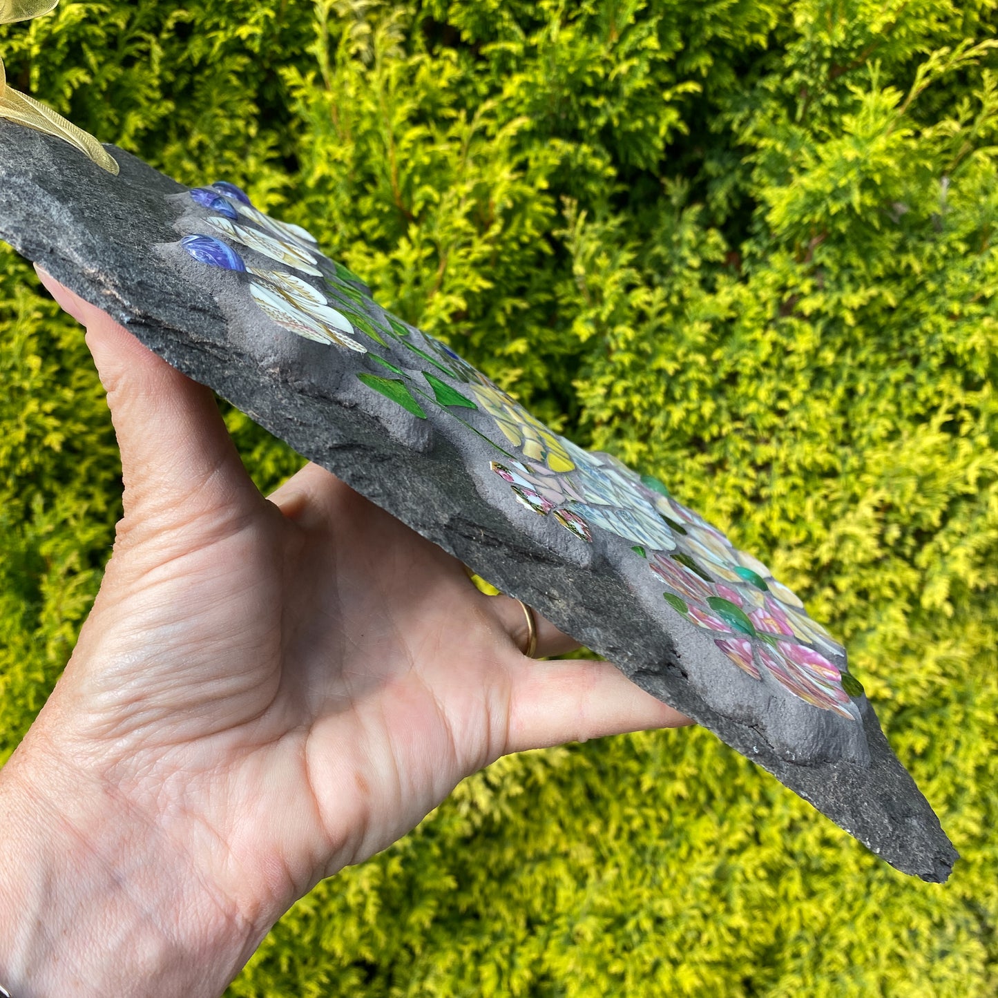 A hand holds a dark slate garden stone decorated with colorful painted lotus flowers, set against a background of bright green foliage.