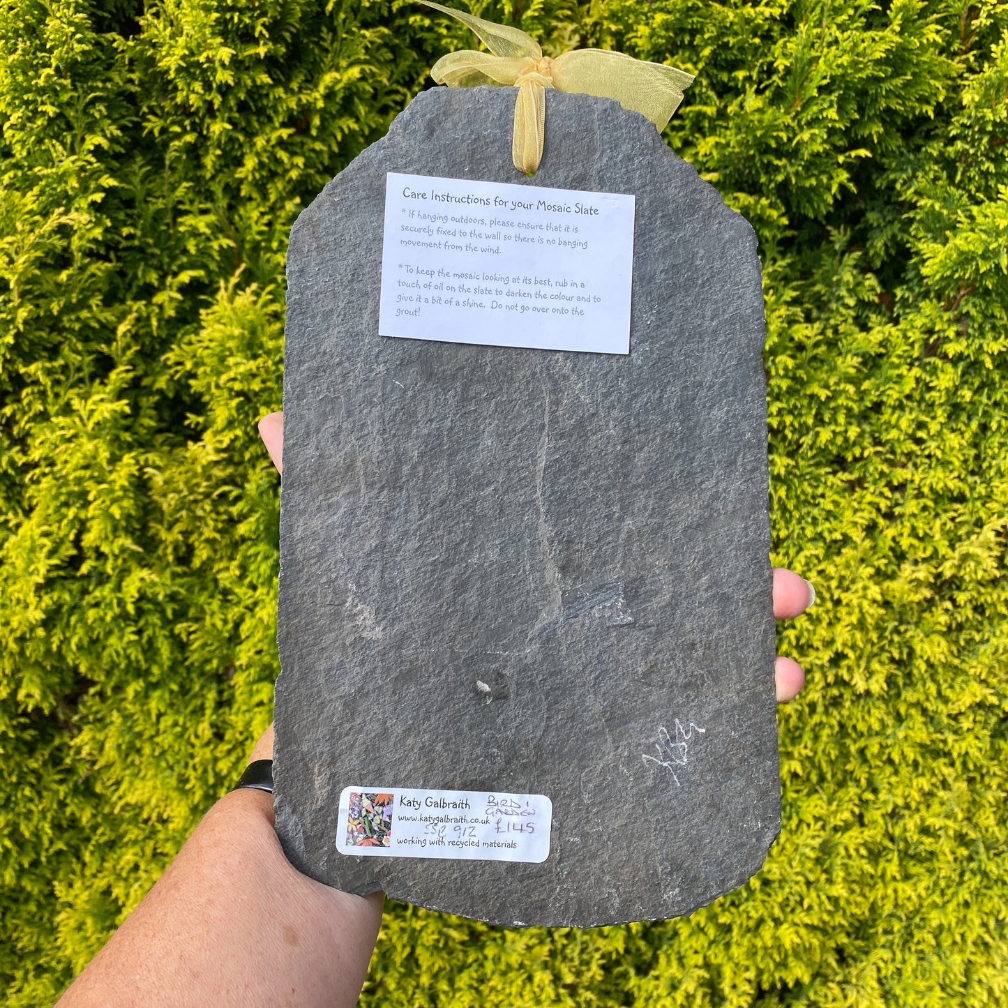 A person holds a dark gray slate with a yellow ribbon bow and two white paper labels, set against a background of bright green foliage.