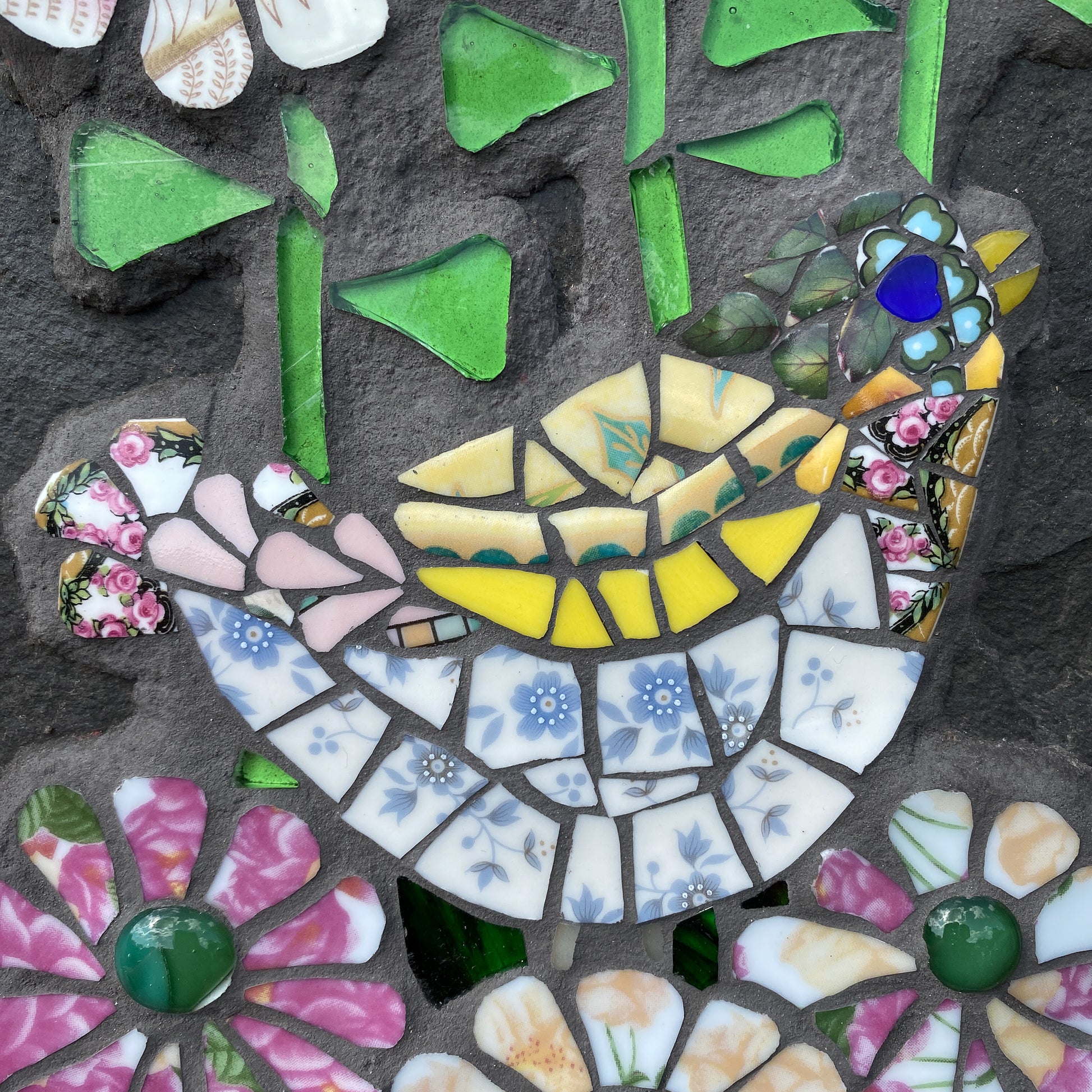 A close-up of a mosaic wall art piece featuring a stylized bird and floral motifs. The design uses broken ceramic tiles in green, yellow, pink, blue, white, and beige, set against a dark textured stone background.