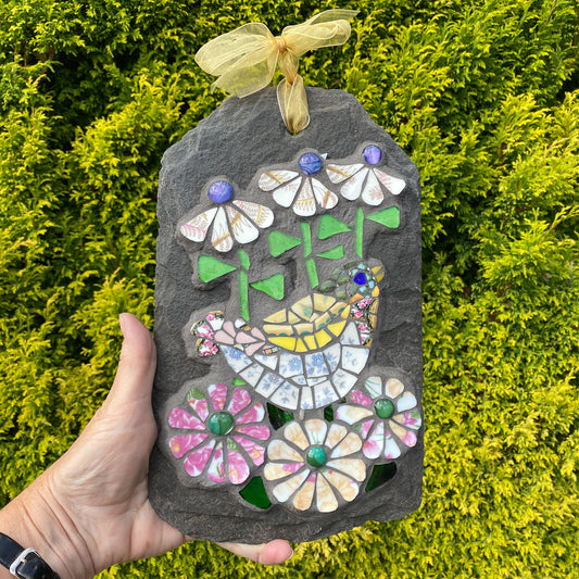 A hand holds a decorative mosaic garden stone ornament featuring a bird and flowers, set against a background of green foliage.